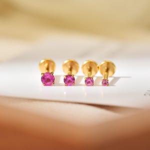 May include: Four gold stud earrings arranged in a row, each featuring a round, vibrant pink gemstone held by four prongs. The earrings are graduated in size, with the largest on the left and the smallest on the right, showcasing a polished finish.