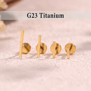 May include: Four gold-coloured G23 Titanium stud earrings. Each earring features a small, round backing with a straight bar design. The earrings are displayed on a textured, off-white surface.