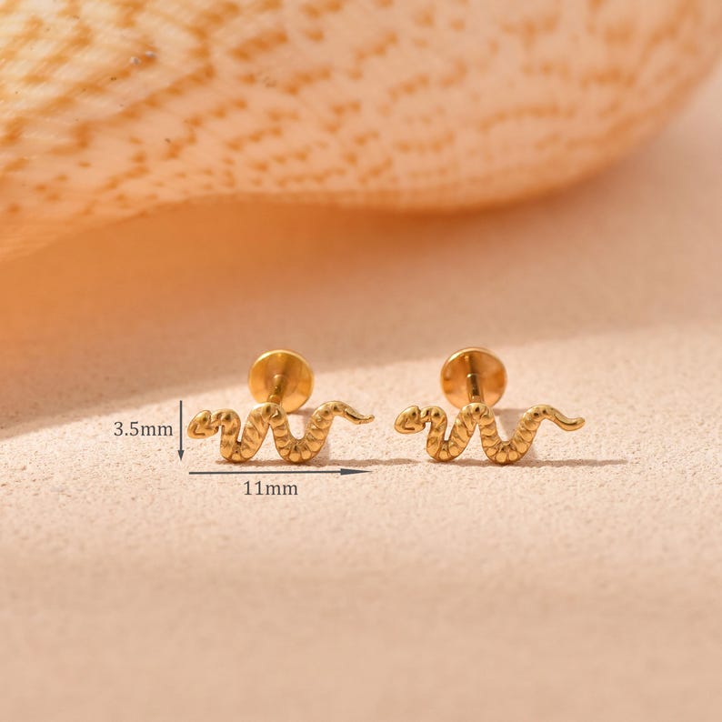 K&ouml;nnte beinhalten: Goldfarbene Schlangen-Ohrstecker. Jeder Ohrring ist 11 mm lang und 3,5 mm breit. Die Ohrringe haben einen Steckerverschluss. Die Ohrringe liegen auf einer hellbraunen Oberfl&auml;che.