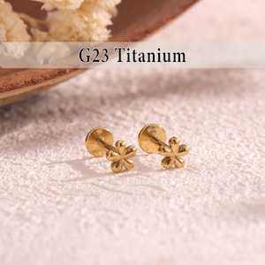May include: Two gold-colored flower-shaped earrings. The earrings have a post and a flat back. The text "G23 Titanium" is displayed above the earrings. The earrings are on a textured, off-white surface.
