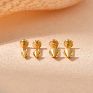 May include: Four gold-toned stud earrings, each featuring a flat circular back and a pointed conical front. The earrings are arranged in a horizontal row, showcasing a polished metallic finish and a simple, geometric spike design.