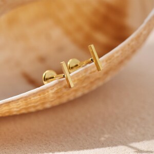 May include: A pair of gold-colored bar stud earrings. The earrings are displayed on a light brown, textured surface. The earrings have a simple, minimalist design, with a small, round stud and a rectangular bar.