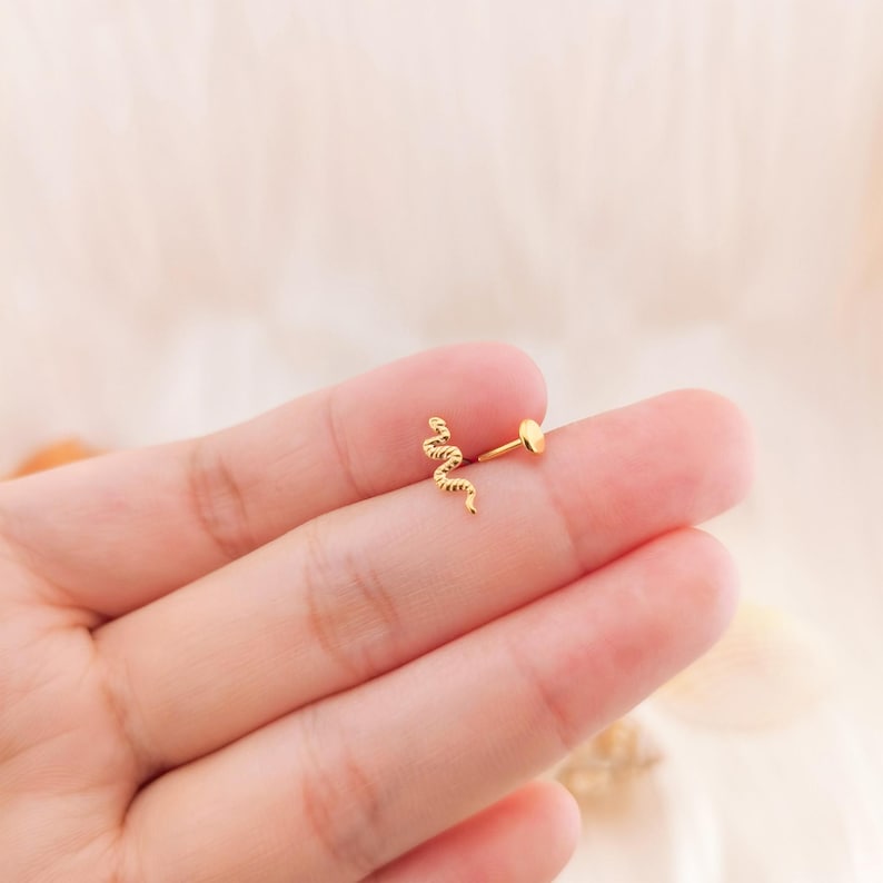 K&ouml;nnte beinhalten: Ein goldfarbener Ohrstecker mit einem kleinen, detaillierten Schlangendesign und einem flachen Scheibenr&uuml;cken. Die Schlange schmiegt sich an das Ohr an und verleiht ihm eine einzigartige und stilvolle Note. Der Schmuck wird auf dem Finger einer Person gezeigt.