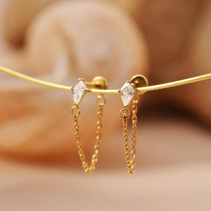 May include: A pair of gold-colored stud earrings with a diamond-shaped, clear gemstone and a dangling chain. The earrings are displayed on a thin, gold-colored wire against a blurred background.