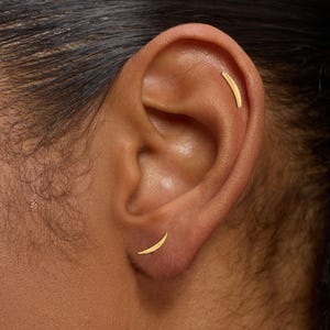 May include: Close-up of an ear adorned with two gold earrings. One earring is a curved bar in the upper ear, and the other is a crescent moon shape on the earlobe. The earrings are a warm gold color.