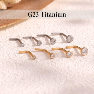May include: An assortment of nose studs in silver and gold tones, each featuring a small, clear gemstone. The text "G23 Titanium" is displayed at the top. The studs are arranged on a textured, off-white surface.