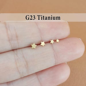 May include: Four small, gold-colored star-shaped earrings are displayed on a person's hand. The earrings are arranged in a row, decreasing in size. The text "G23 Titanium" is displayed above the earrings.