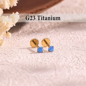 May include: A pair of gold-coloured G23 Titanium earrings, each featuring a blue opal-like stone. The earrings have a flat disc backing and a post with a small, round, blue stone. The text "G23 Titanium" is displayed above the earrings.