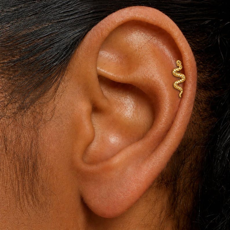 K&ouml;nnte beinhalten: Ein goldfarbener Ohrring in Schlangenform ist am oberen Ohr zu sehen. Der Ohrring ist ein kleines, gebogenes Design mit einer strukturierten Oberfl&auml;che, die einer Schlange &auml;hnelt. Der Ohrring ist vor dem Hintergrund des nat&uuml;rlichen Hauttons des Ohrs platziert.