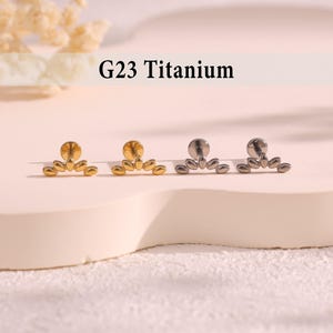May include: Four pieces of jewellery are displayed. Two are gold-toned, and two are silver-toned. Each piece has a flat, round base with a decorative, fan-shaped design. The text "G23 Titanium" is above the jewellery.