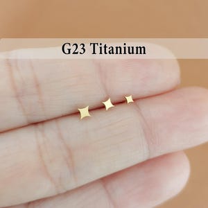 May include: Three small, gold-colored star-shaped earrings. The earrings are arranged in a row, decreasing in size. The text "G23 Titanium" is displayed above the earrings.