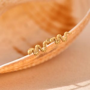 K&ouml;nnte beinhalten: Ein goldfarbener Ohrring in Schlangenform mit strukturierter Oberfl&auml;che. Der Ohrring befindet sich auf einer hellen, gebogenen Oberfl&auml;che, m&ouml;glicherweise einer Muschel. Der Hintergrund ist in einem weichen, warmen Pfirsichton gehalten.