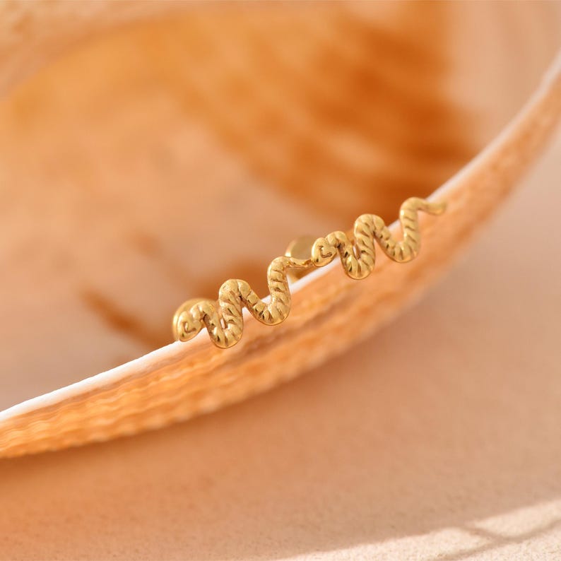 K&ouml;nnte beinhalten: Ein goldfarbener Ohrring in Schlangenform mit strukturierter Oberfl&auml;che. Der Ohrring befindet sich auf einer hellen, gebogenen Oberfl&auml;che, m&ouml;glicherweise einer Muschel. Der Hintergrund ist in einem weichen, warmen Pfirsichton gehalten.