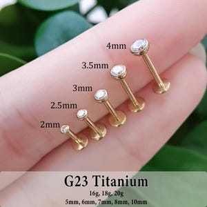 May include: Five gold-colored G23 titanium labret studs with clear round gemstones, ranging in size from 2mm to 4mm. The text G23 Titanium, 16g, 18g, 20g, and 5mm, 6mm, 7mm, 8mm, 10mm is printed at the bottom of the image for reference.