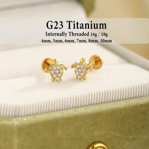 May include: Two gold-tone turtle stud earrings with clear stones, featuring G23 Titanium, internally threaded 16g/18g, and available in 4mm, 5mm, 6mm, 7mm, 8mm, and 10mm lengths. The turtle shells are encrusted with small, sparkling white stones.