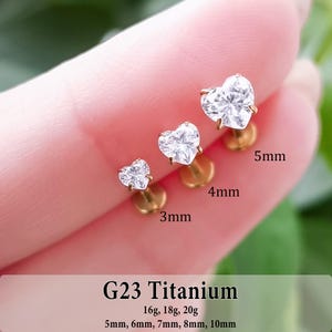May include: Three gold-tone G23 titanium labret studs with heart-shaped clear cubic zirconia stones. The studs are labeled with sizes 3mm, 4mm, and 5mm. Text at the bottom indicates G23 Titanium, gauges 16g, 18g, 20g, and lengths 5mm to 10mm.