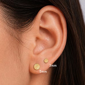 May include: Close-up of two gold stud earrings, one 5mm and the other 3mm in size, each with a sunburst design. The earrings are displayed on an ear, showcasing their size and style. The image highlights the jewelry's simple elegance.