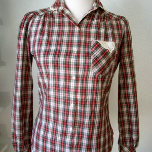 Puede incluir: Una camisa de manga larga a cuadros rojos, blancos y verdes con un cuello y puños de encaje blanco. La camisa tiene un solo bolsillo en el pecho izquierdo con un borde de encaje blanco.