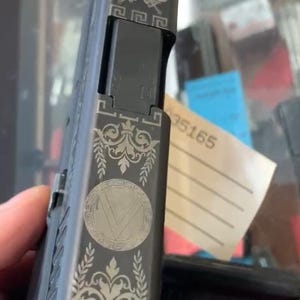Puede incluir: Una pistola negra con un diseño grabado en plata que presenta una espada, espadas cruzadas y un patrón floral. El grabado también incluye un borde de clave griega y un diseño circular con una letra V en el centro.
