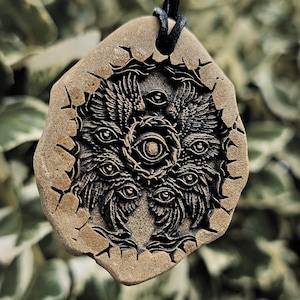 May include: A tan, irregularly shaped stone pendant suspended on a black cord. The surface features a deeply engraved, dark, intricate design of a multi-eyed, winged entity surrounded by a jagged, cracked circular border with radiating lines.