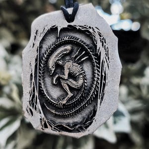 May include: A grey, irregularly shaped pendant featuring a detailed, embossed relief of a xenomorph creature in a curled position. The pendant is suspended from a thin black cord, showcasing a dark, recessed design against a lighter grey surface.