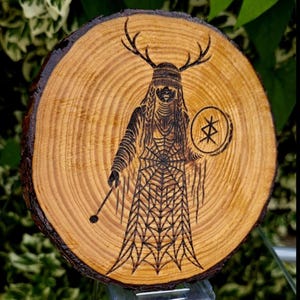 Peut inclure: Une tranche de bois circulaire présentant une gravure au pyrographe représentant une silhouette à bois de cerf, vêtue d'une longue robe à motif de toile d'araignée. La silhouette tient un bouclier circulaire avec un symbole étoilé et un bâton.
