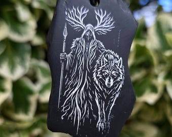 May include: A black stone pendant featuring a white engraved illustration of a hooded figure with large antlers holding a spear, standing beside a wolf. The pendant is suspended from a thin black cord and has an irregular, natural edge shape.