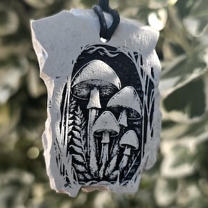 May include: A pendant featuring a black and white relief print of a cluster of mushrooms and ferns on a textured, irregular off-white stone-like material, suspended from a thin black cord for wear as a necklace or decorative hanging ornament.