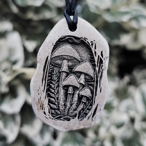May include: A grey, irregularly shaped stone pendant featuring a detailed, dark, recessed engraving of a cluster of four mushrooms surrounded by ferns. The pendant is suspended from a simple black cord, creating a contrast between the light stone and the dark, intricate mushroom illustration.