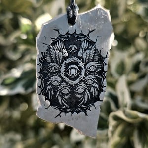 Op de afbeelding: Een hanger van onregelmatig, gebroken wit, steenachtig materiaal met een zwarte inktillustratie van een cirkelvormige, gevleugelde entiteit met meerdere ogen. De hanger hangt aan een dun zwart koord en toont gedetailleerd lijnwerk.