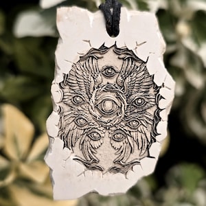 May include: A rectangular off-white stone pendant featuring a detailed dark engraving of a multi-eyed, winged celestial being surrounded by a crown of thorns. The pendant has a rough, uneven edge and is suspended from a simple black cord necklace.