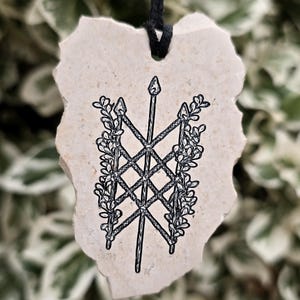 May include: A beige, irregularly shaped stone pendant suspended by a black cord. The surface features a black ink illustration of a central spear crossed by two others, forming a lattice pattern framed by leafy vines on both sides.