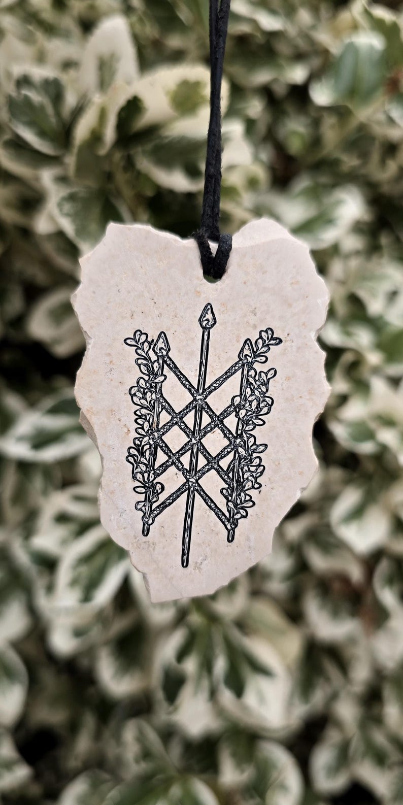 May include: A beige, irregularly shaped stone pendant suspended by a black cord. The surface features a black ink illustration of a central spear crossed by two others, forming a lattice pattern framed by leafy vines on both sides.