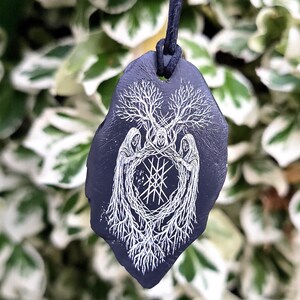 May include: A dark purple, leaf-shaped pendant hanging from a black cord. The pendant features a white, intricate line-art illustration of three hooded figures standing in a circle, surrounded by tree branches and roots with a central geometric symbol.