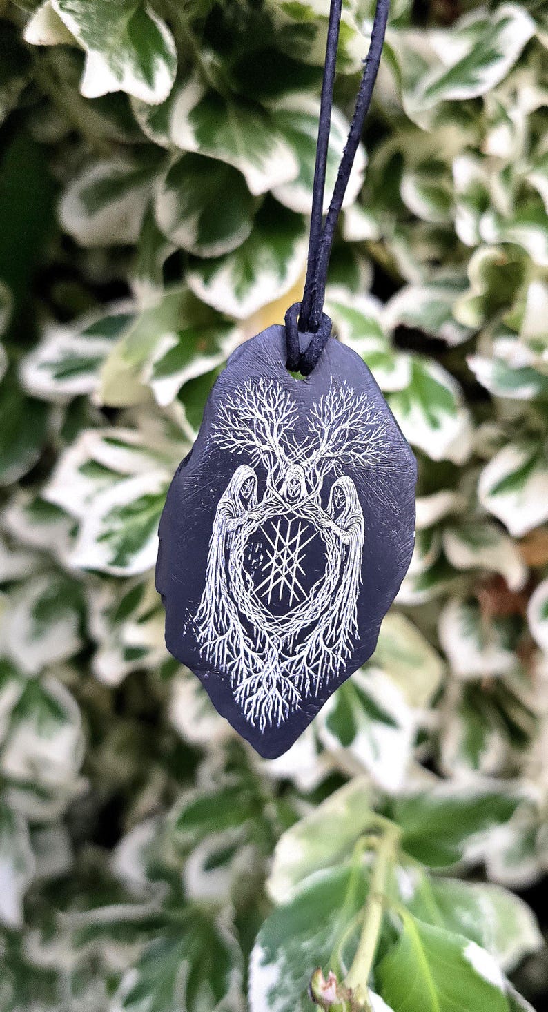 May include: A dark purple, leaf-shaped pendant hanging from a black cord. The pendant features a white, intricate line-art illustration of three hooded figures standing in a circle, surrounded by tree branches and roots with a central geometric symbol.