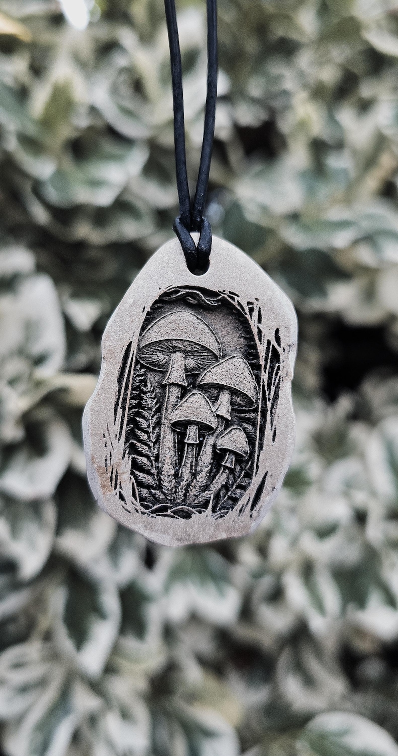 May include: A grey, irregularly shaped stone pendant featuring a detailed, dark, recessed engraving of a cluster of four mushrooms surrounded by ferns. The pendant is suspended from a simple black cord, creating a contrast between the light stone and the dark, intricate mushroom illustration.