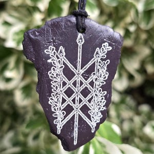 May include: A dark purple, irregularly shaped stone pendant suspended from a black cord. The surface features a white, intricate line-art design of a central spear crossed by two others, flanked by detailed leafy vine patterns on both sides.