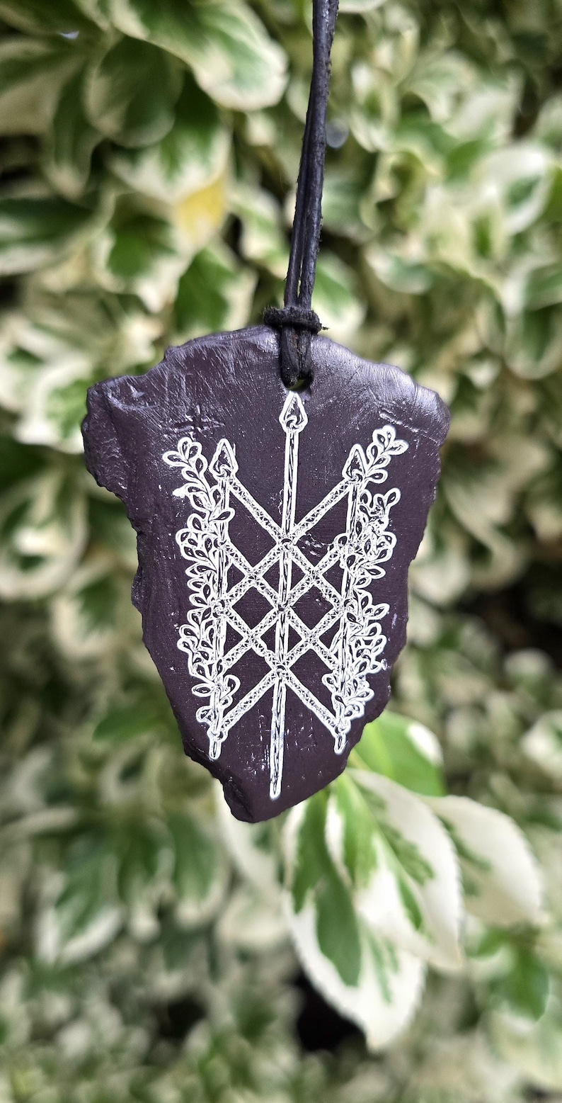 May include: A dark purple, irregularly shaped stone pendant suspended from a black cord. The surface features a white, intricate line-art design of a central spear crossed by two others, flanked by detailed leafy vine patterns on both sides.
