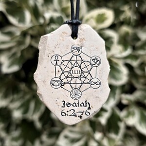 May include: A cream-colored, irregular stone pendant hanging from a black cord. The surface features a black ink print of a geometric Metatron's Cube design with various symbols, the number 1111, and the text Isaiah 6:2-6 printed at the bottom.