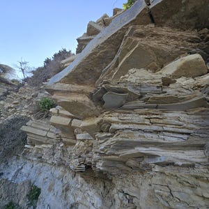 May include: A steep, layered rock cliff face featuring tan and grey sedimentary stone formations. The rugged surface displays horizontal strata and jagged, protruding rock slabs under a clear blue sky with sparse green vegetation.
