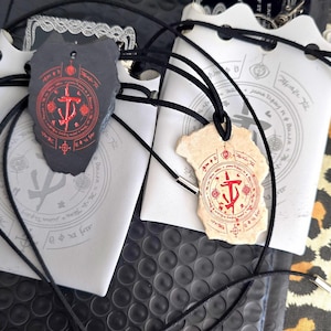 May include: Two irregular stone-like pendants on black cord necklaces. One pendant is dark gray with a red circular symbol, and the other is light beige with a matching red symbol. Both are displayed on white cards featuring circular graphic designs.