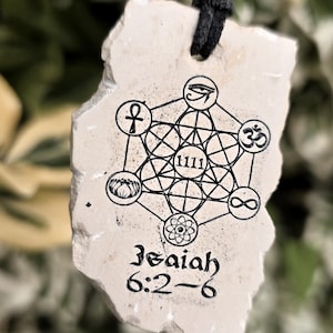 May include: A beige, irregularly shaped stone pendant hanging from a black cord. The surface features a black ink print of a geometric pattern, the number 1111, various symbols, and the text Isaiah 6:2-6 in a stylized black font.