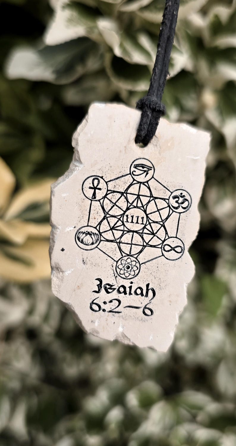 May include: A beige, irregularly shaped stone pendant hanging from a black cord. The surface features a black ink print of a geometric pattern, the number 1111, various symbols, and the text Isaiah 6:2-6 in a stylized black font.