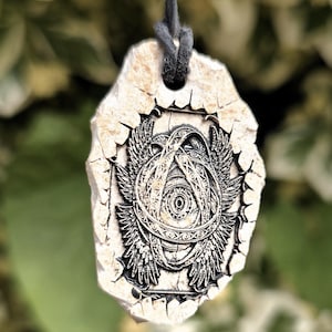 May include: A pendant featuring a detailed, dark-toned engraving of a winged eye surrounded by concentric rings, set into a textured, off-white, irregular stone-like material. The pendant hangs from a thin, dark grey cord through a top hole.