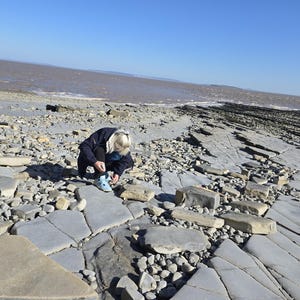 May include: A person wearing a dark blue jacket and light blue sneakers crouches on a rocky beach with flat grey stone slabs and pebbles, with a view of the brown ocean water and a clear blue sky in the background on a sunny day.
