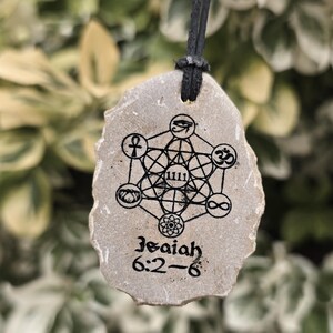 May include: A tan, irregularly shaped stone pendant hanging from a black cord. The stone features a black ink print of a geometric Metatron's Cube design with various symbols, the number 1111, and the text Isaiah 6:2-6 printed at the bottom.