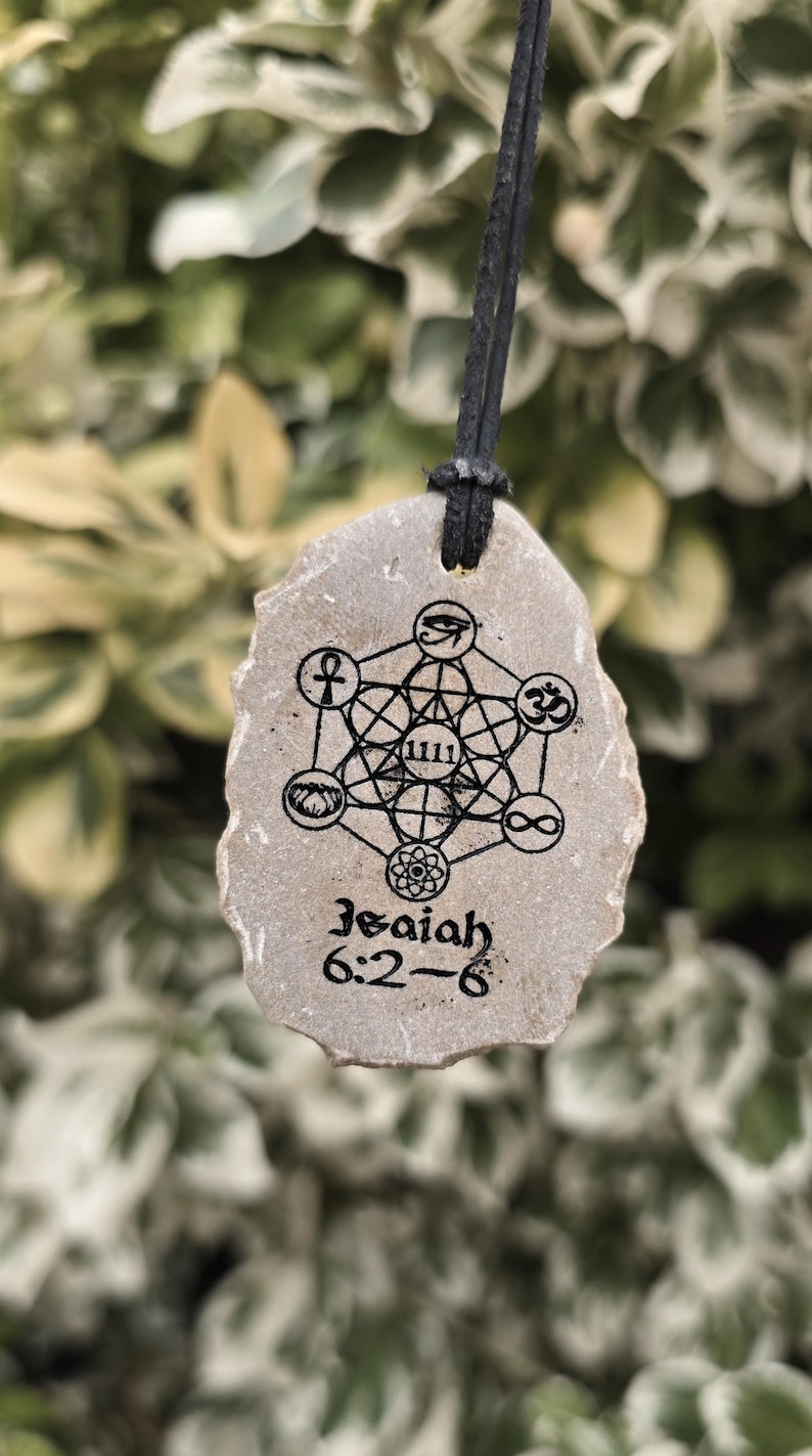 May include: A tan, irregularly shaped stone pendant hanging from a black cord. The stone features a black ink print of a geometric Metatron's Cube design with various symbols, the number 1111, and the text Isaiah 6:2-6 printed at the bottom.