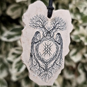 May include: A cream-colored, irregular-shaped stone pendant hanging from a black cord. The surface features a black ink illustration of three hooded figures standing in a circle, surrounded by tree branches and roots, with a geometric symbol in the center.