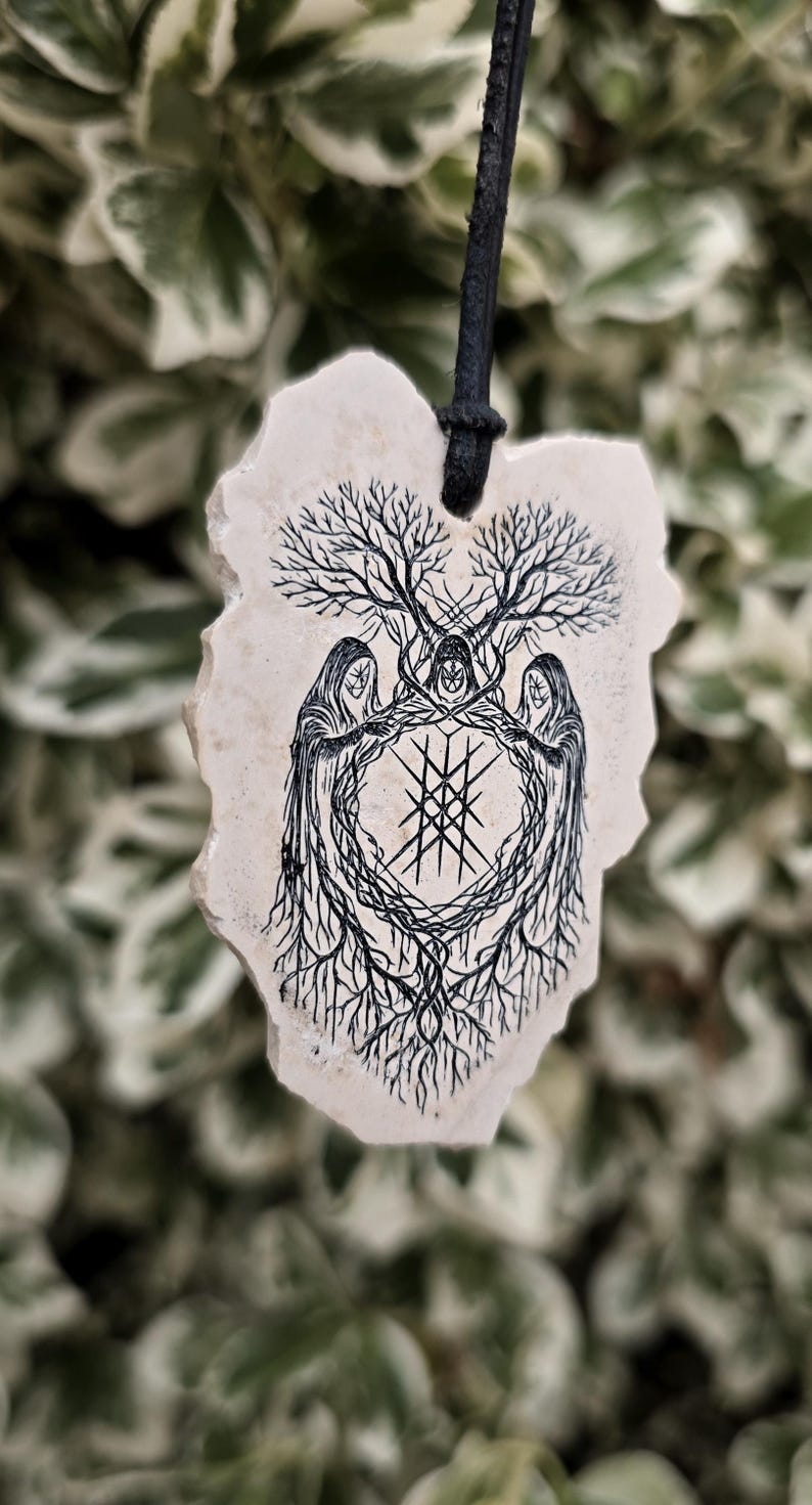May include: A cream-colored, irregular-shaped stone pendant hanging from a black cord. The surface features a black ink illustration of three hooded figures standing in a circle, surrounded by tree branches and roots, with a geometric symbol in the center.