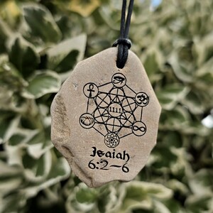 May include: A tan, irregularly shaped stone pendant hanging from a black cord. The stone features a black printed design of Metatron's Cube with various symbols, the number 1111, and the text Isaiah 6:2-6 printed in a black serif font.